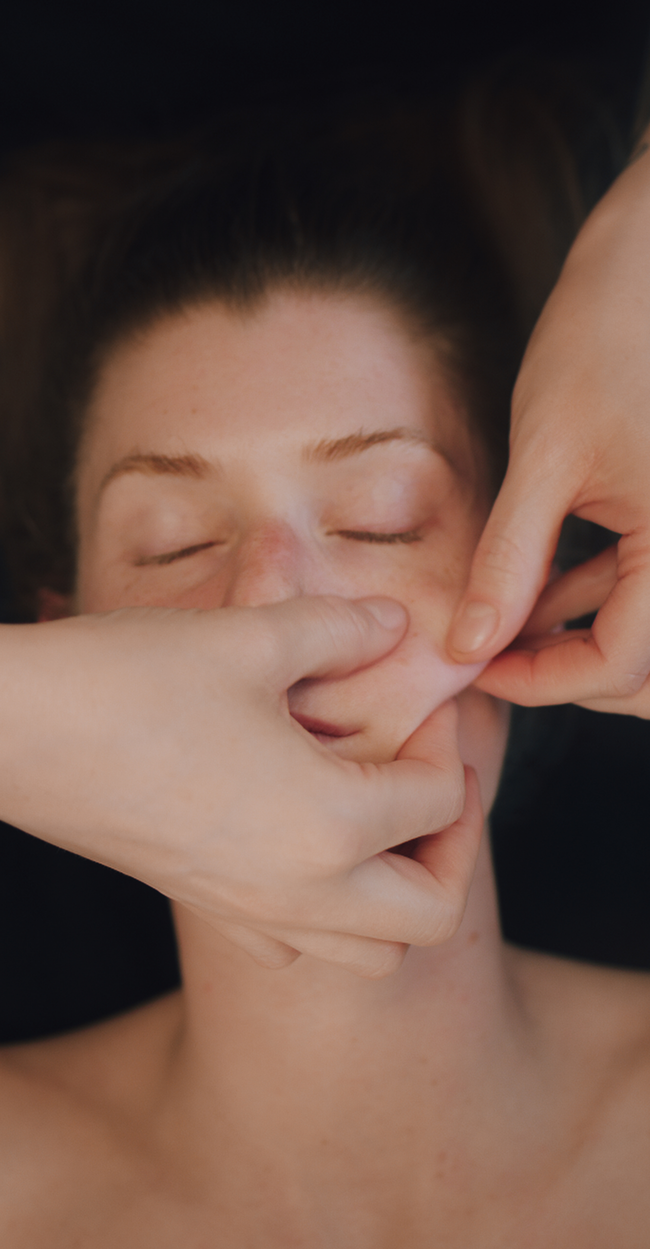 Eine Person mit geschlossenen Augen erhält eine Gesichtsmassage, während zwei Hände sanft ihre Wangen drücken und kneten, was auf Entspannung und Hautpflege hindeutet.